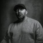 Black and white portrait of a man in a casual sweater and cap.